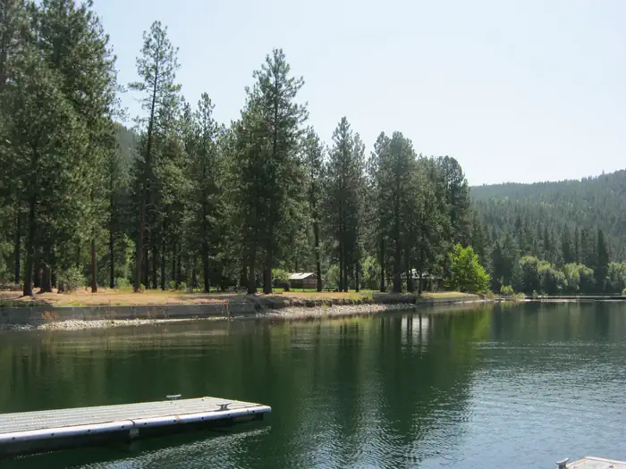 Evans Campground — The photo shows a calm lakeshore lined with tall pines and a small dock in the foreground, with forested hills rising in the background.