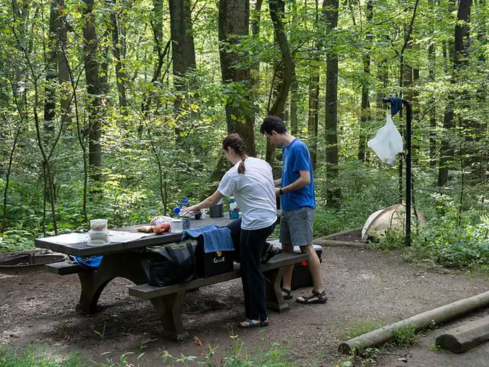 Owens Creek Campground — Man and woman standing next to picnic table on wood chip campsite. On the tent pad behind the campers is a tent. Waste back hangs from lantern pole. Site is surrounded by dense forest.