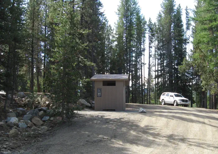 Olive Lake Campground (or) — brown vault toilet beside gravel road with white SUV next to pine trees