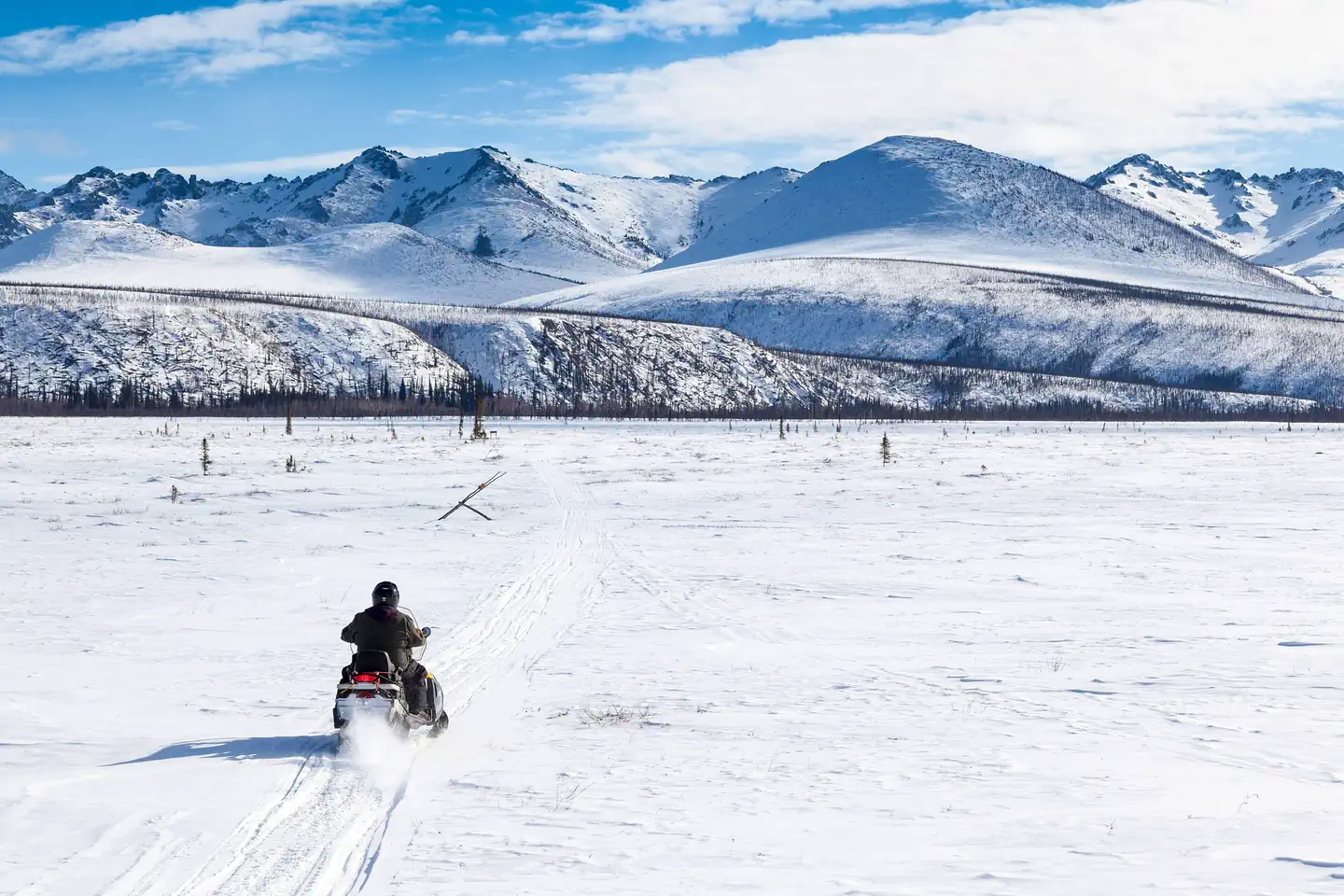 White Mountains National Recreation Area - Alaska Cabins — A snowmobile riders crosses a big meadow with mountains in the distance.