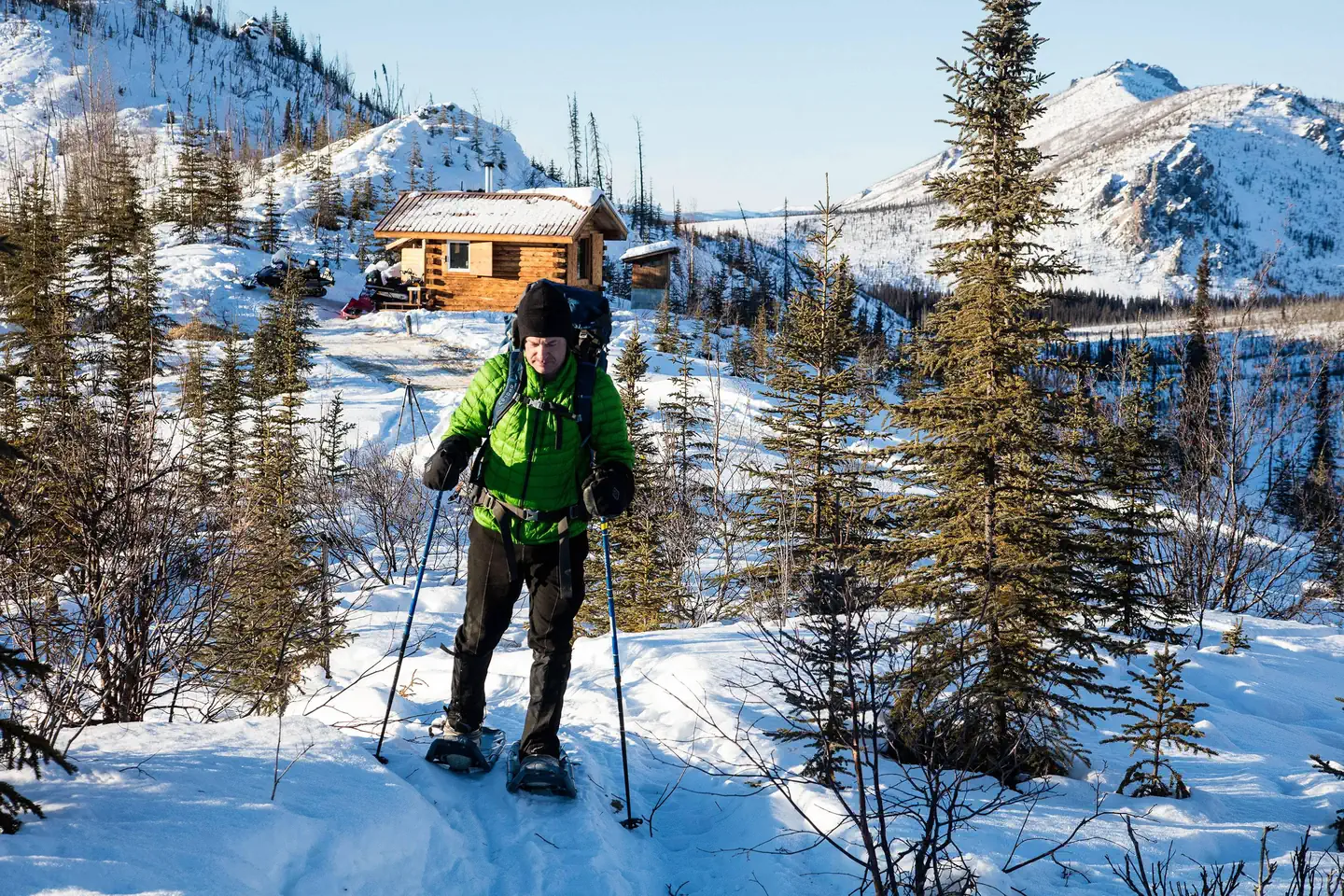 White Mountains National Recreation Area - Alaska Cabins — A man snowshoes from a log cabin