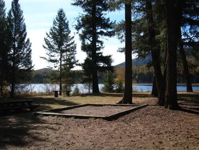 Lost Lake Group Unit — Sunlit tent pad in front of conifers in silhouette with lake and hill in background.