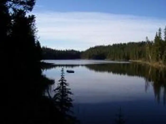 Swan Lake Kitchen β Glassy lake reflecting conifer lined shore, blue sky and cloud.