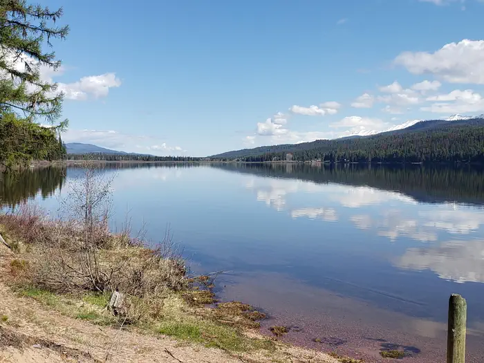 Seeley Lake Campground — View from Seeley Lake beach looking North. A deep, blue sky and trees lining the lake can be seen reflecting in the water.