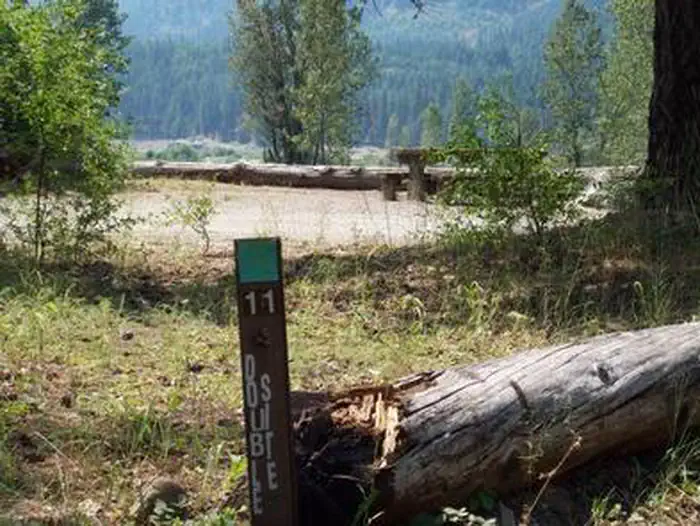 Cle Elum River Campground — Site marker eleven in foreground; picnic table in a flat gravel area with bushes encircled with log barrier, river valley and conifer covered hills in background.