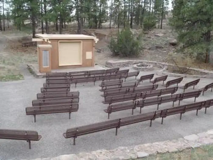 Juniper Family Campground — Amphitheater with brown benches and a tan building surrounded by pine trees