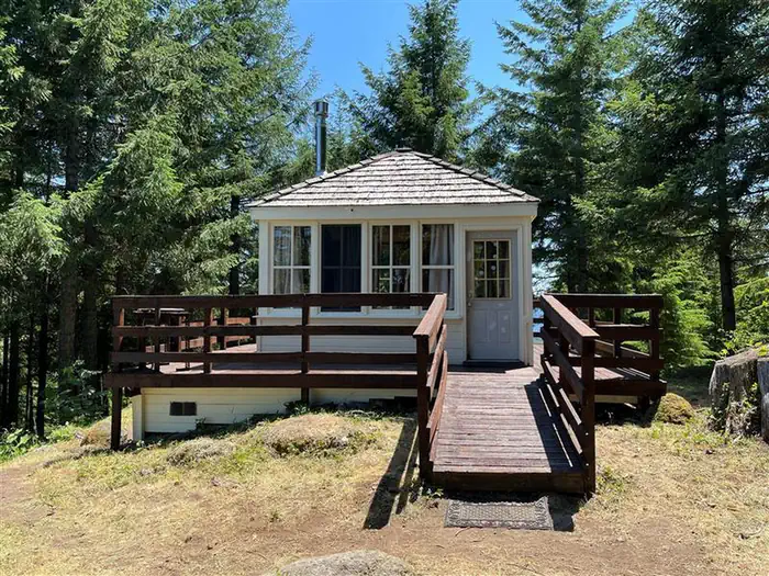 Timber Butte Cabin — Short ramp leading onto wrap around porch of white cabin, fir trees surround the cabin.