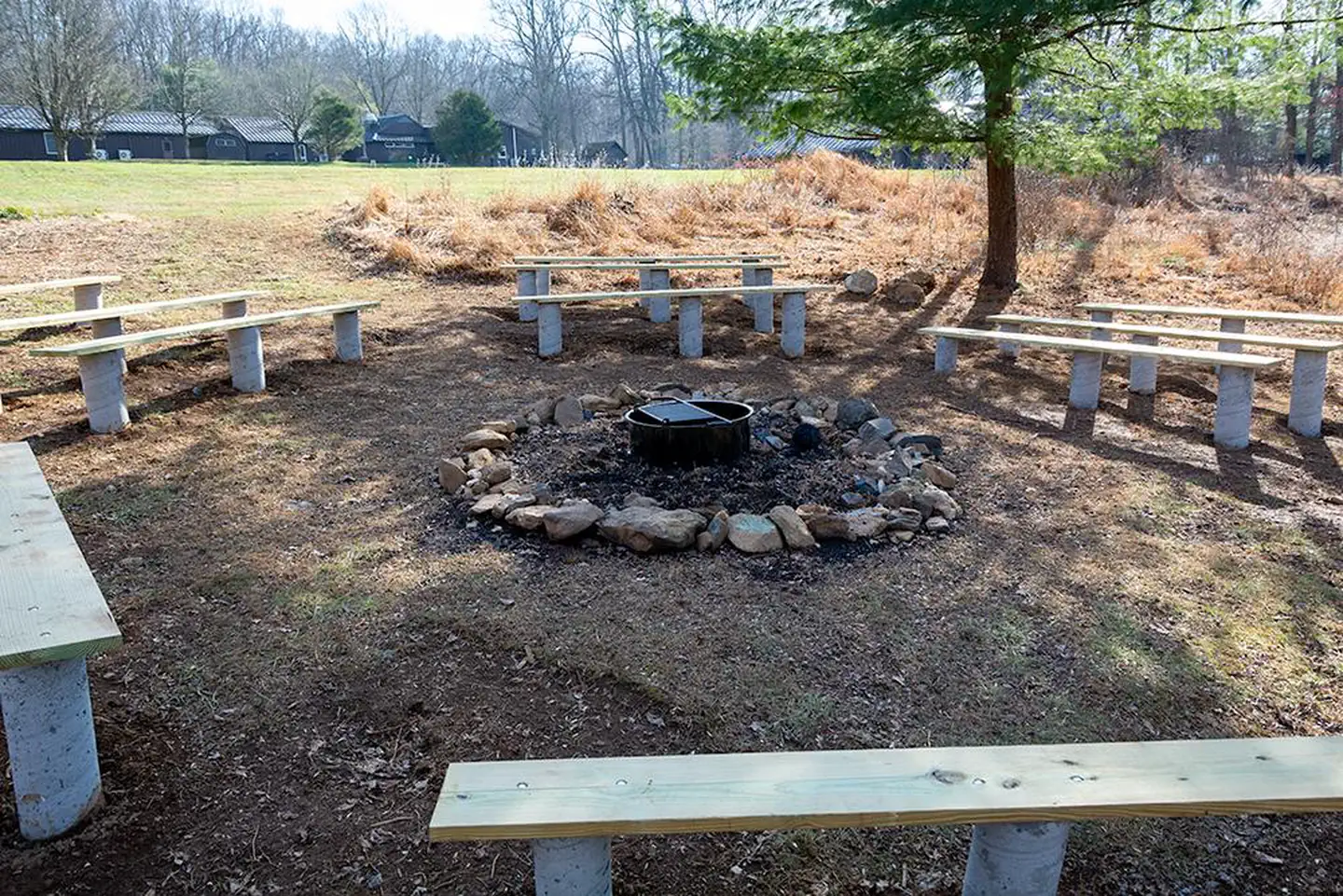 Camp Round Meadow — Outdoor fire circle located below athletic field. Metal fire ring/grill surrounded by a stones. Wooden benches on concrete pilings. Rear of dining hall in the background