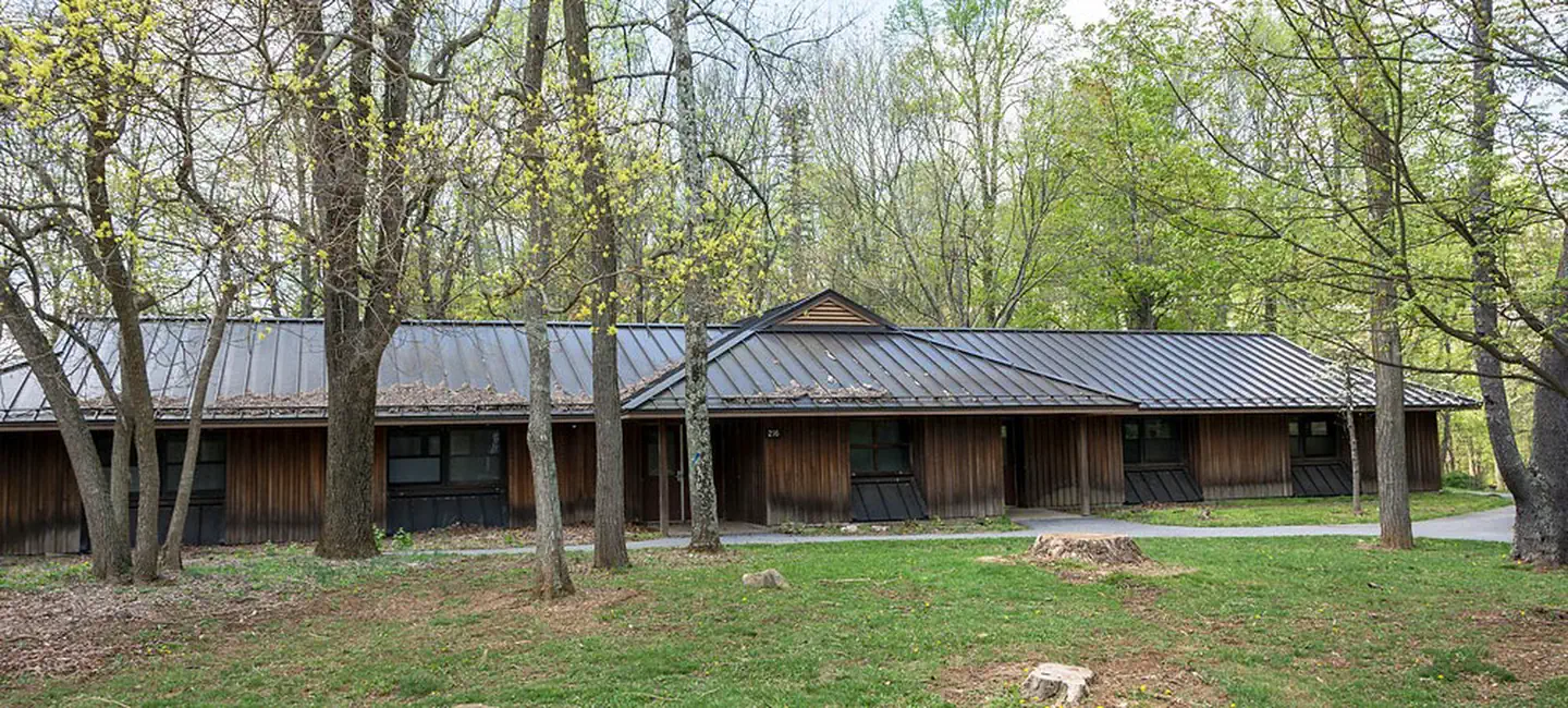 Camp Round Meadow — Front view of one dormitory building in early spring. Large trees are visible in front and behind the dormitory. Each dorm accommodates 30 people and is heated.