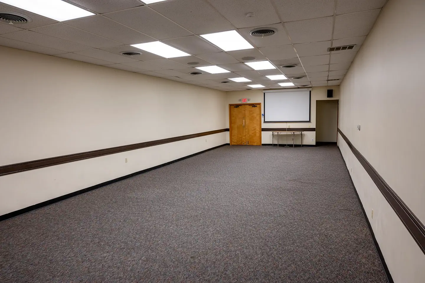 Camp Round Meadow — Interior view of the small conference room. Gray carpet flooring with 2 doors at the far end. Ceiling mounted video screen on far end of room.