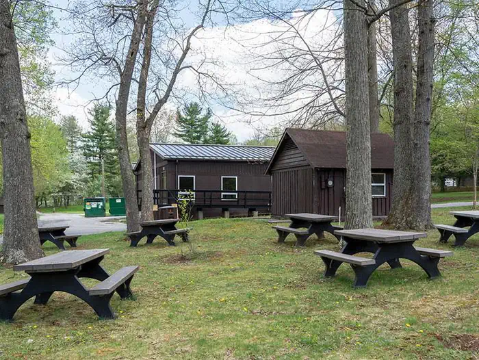Camp Round Meadow — Multiple picnic tables outdoors in grass area on the side of the dining hall facility. Several large trees shade the picnic area.