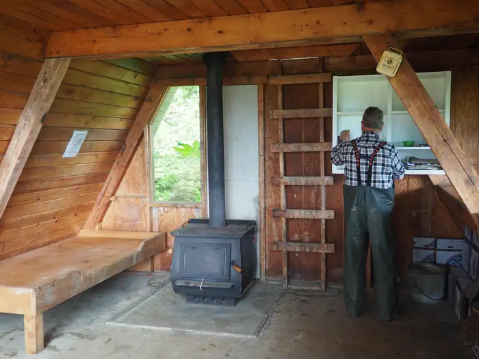 Little Dry Island Cabin — Man at shelves inside wood cabin with black stove and wood bunk