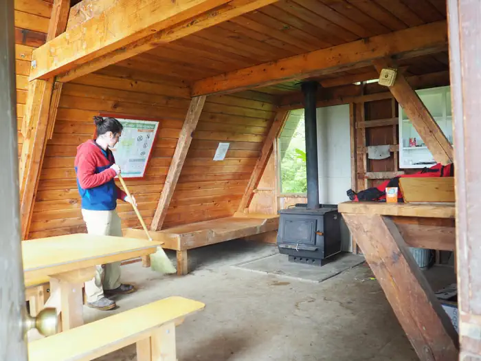 Little Dry Island Cabin — Person sweeping the interior of wood cabin with wood table and bunkbed with a black stove