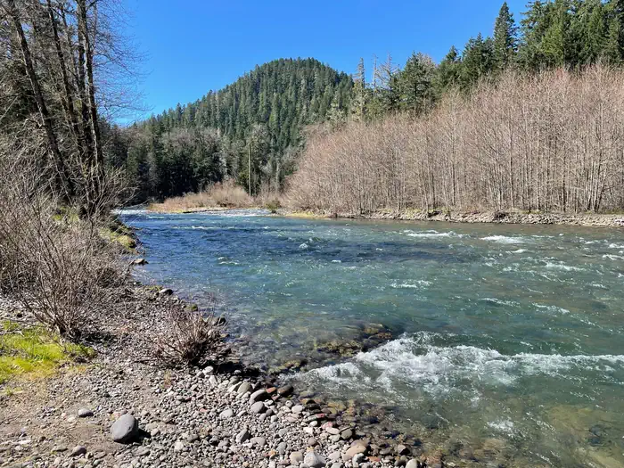 Sand Prairie Campground — A rocky beach leading into a blue river. Small tree covered peaks can be seen on the other bank.