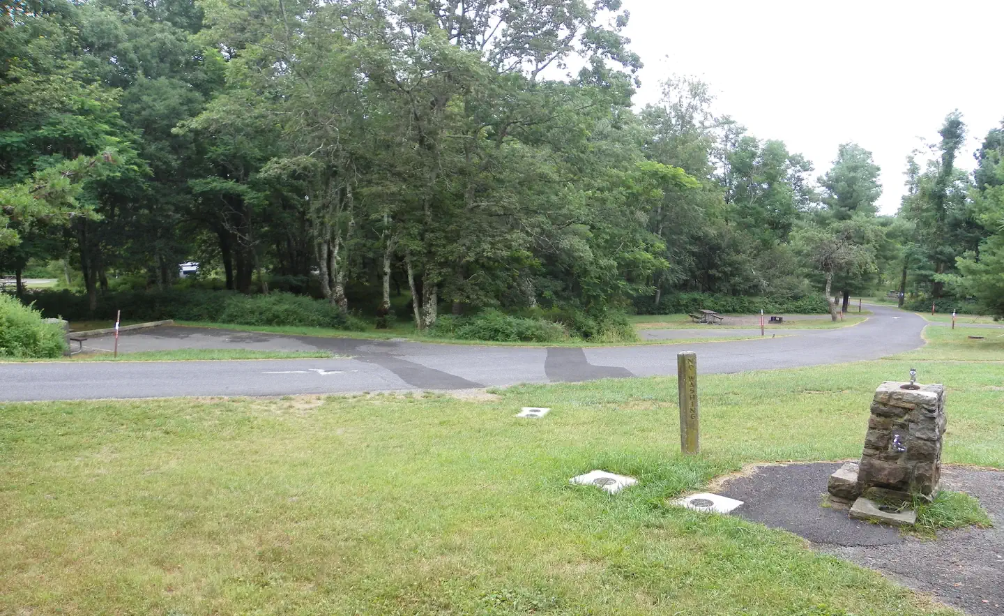 Big Meadows Campground - Shenandoah (va) — A stone water fountain next to a road that is lined with campsites. Each campsite is surrounded by trees and bushes.