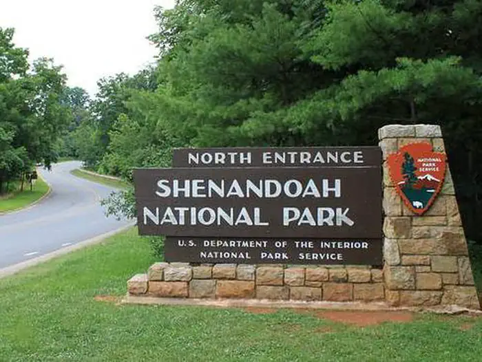 Big Meadows Campground - Shenandoah (va) — A narrow road winds between trees, past a stone post holding a wooden sign that reads "North Entrance Shenandoah National Park" with the National Park service arrowhead logo.