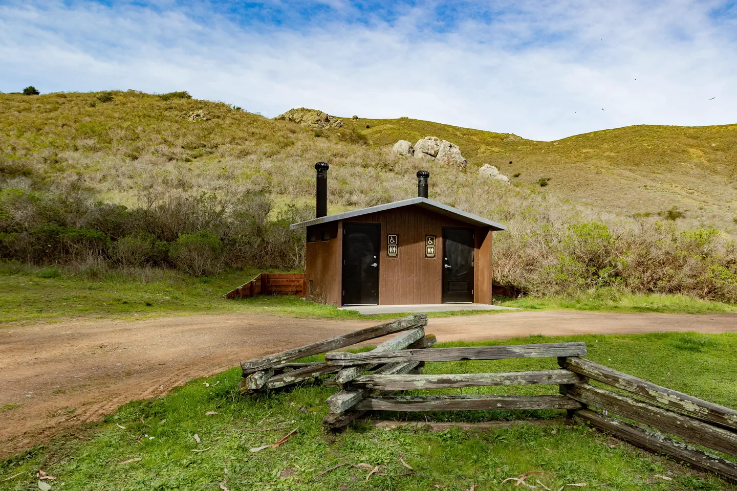 Haypress Campground — A pit toilet structure sits in front of along a dirt path. In the distance are shrub covered hills.