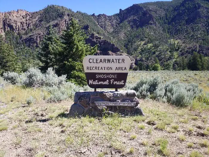 CLEARWATER CAMPGROUND — Yellow and brown campsite sign mounted on a rock pedestal. Trees, mountains and a little bit of blue sky are in the background.