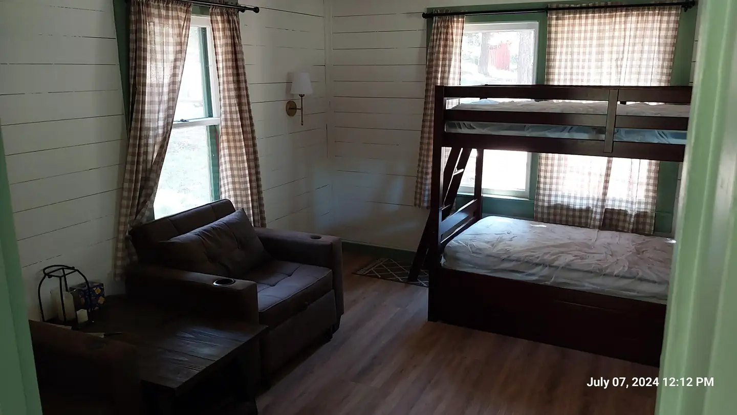 Patterson Guard Station — Bunk beds and chairs in the main living room seen from the kitchen. The scene is dimly lit as sunlight enters the room through 3 large windows.