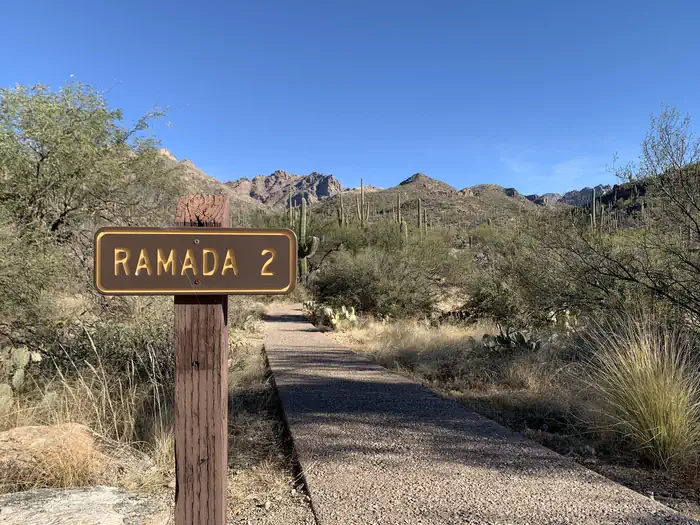 SABINO CANYON RECREATION AREA CACTUS RAMADA 2 — Brown sign to the left of a paved path that says "Ramada 2"