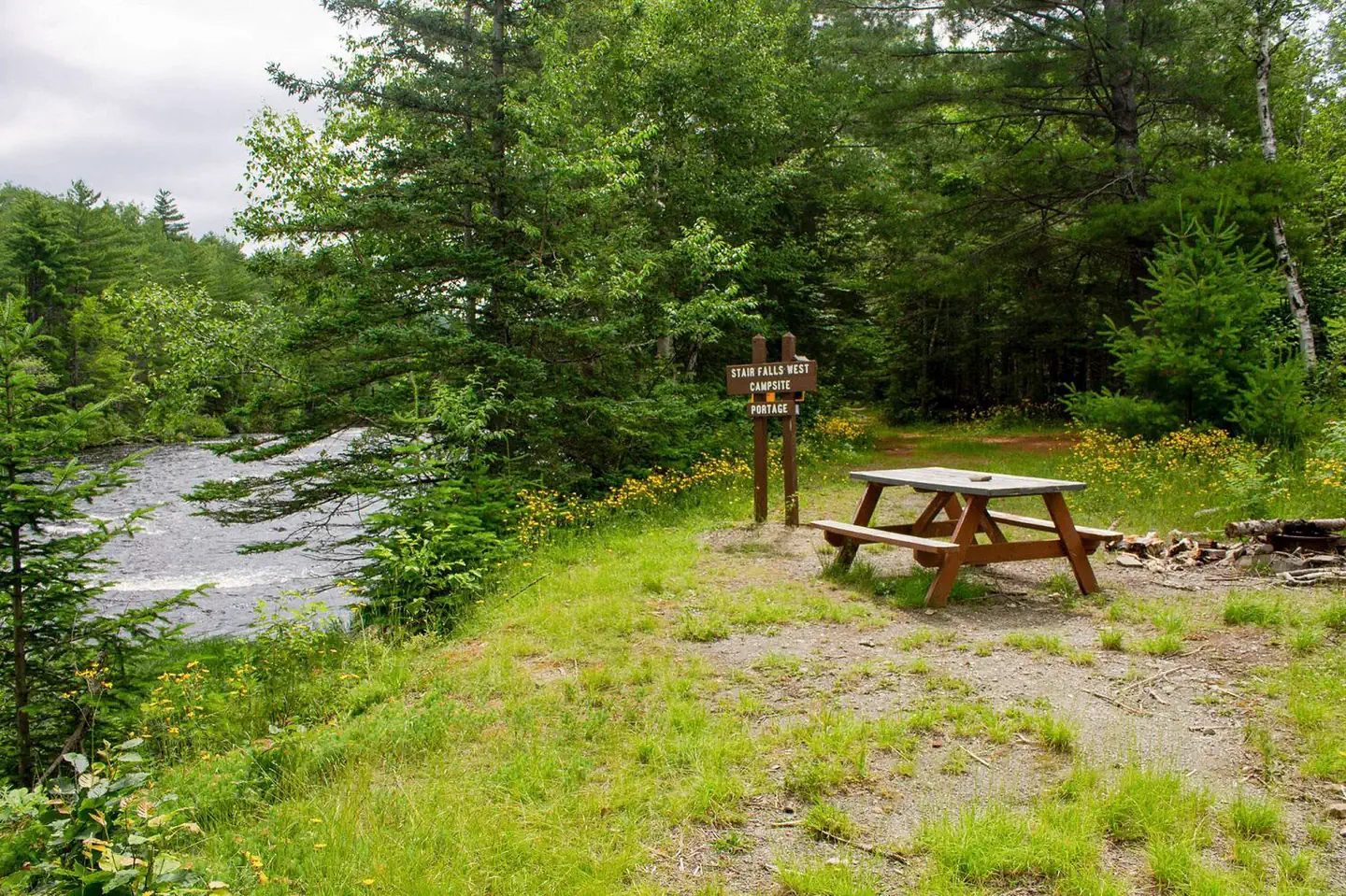Katahdin Woods & Waters National Monument Camping — A gravel and grassy campsite to the right of a river. The campsite has a wooden picnic table, a brown wooden sign, and a trail that leads into the woods with tall dense green trees.