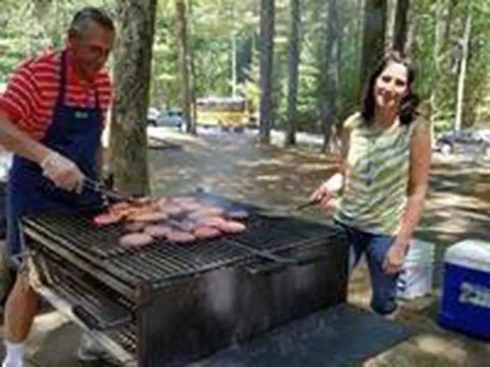WEST HILL PARK β View of two adults cooking on charcoal grill (on site provided) with coolers, parking lot and parked cars in the background.