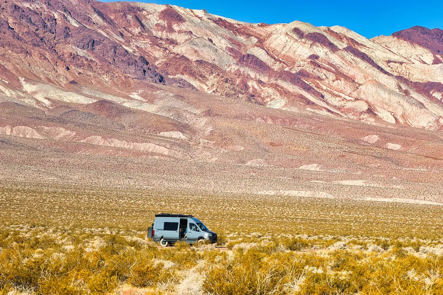 Death Valley Backcountry Roadside Camping — Gray campervan on roadside campsite with green brush vegetation in the foreground and dramatic reddish and white stratified mountains in the far background.