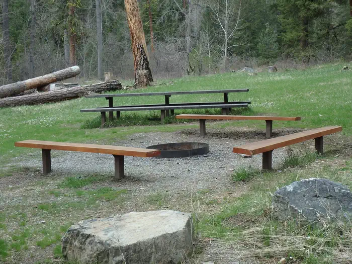 Pine Flats Group Campground — Large flat rock in front of benches around a fire ring with picnic table, grassy meadow, and forest in background.