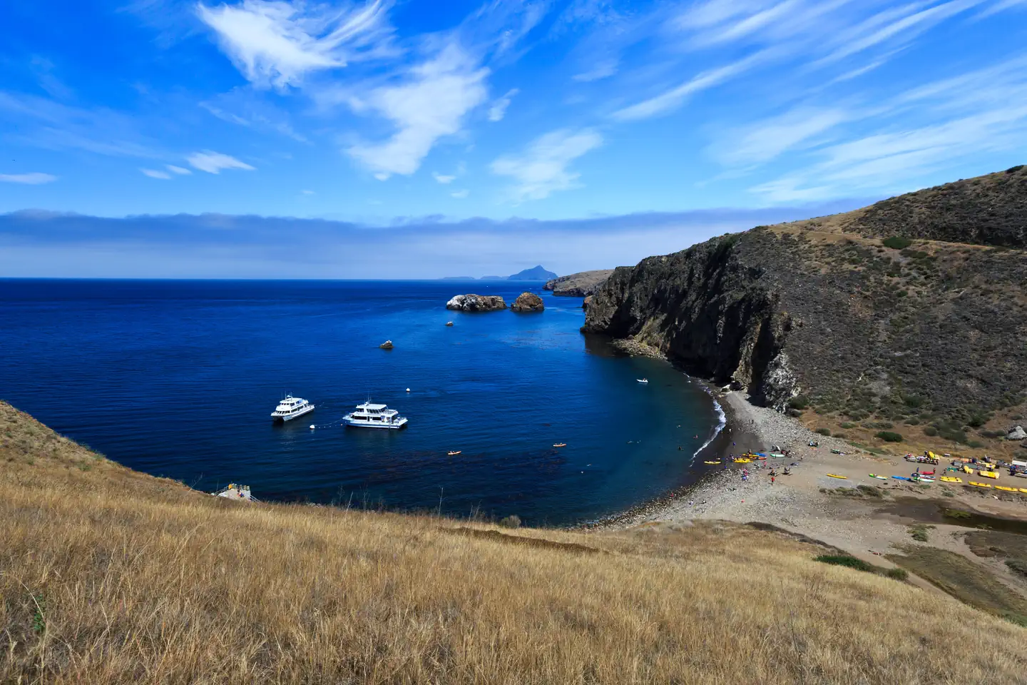 Santa Cruz Island Scorpion Canyon Campground — Harbor with boats, kayaks, and visitors on the beach. Steep island cliffs and other islands in the distance. 