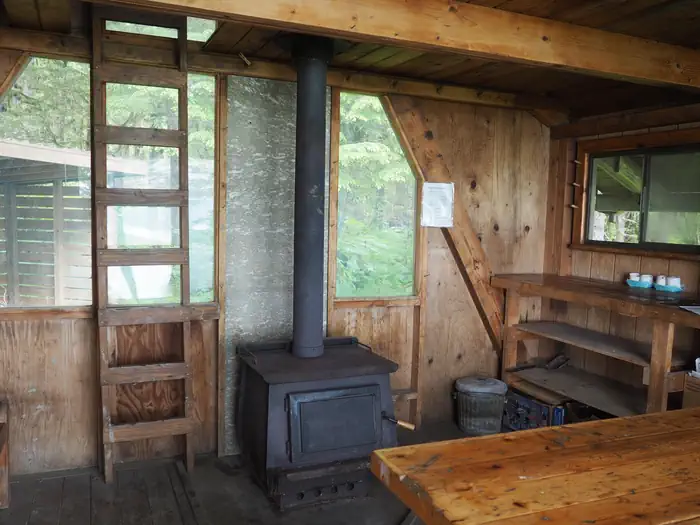 Mallard Slough Cabin — Interior of wood cabin with shelves, tables, stove and ladder to loft