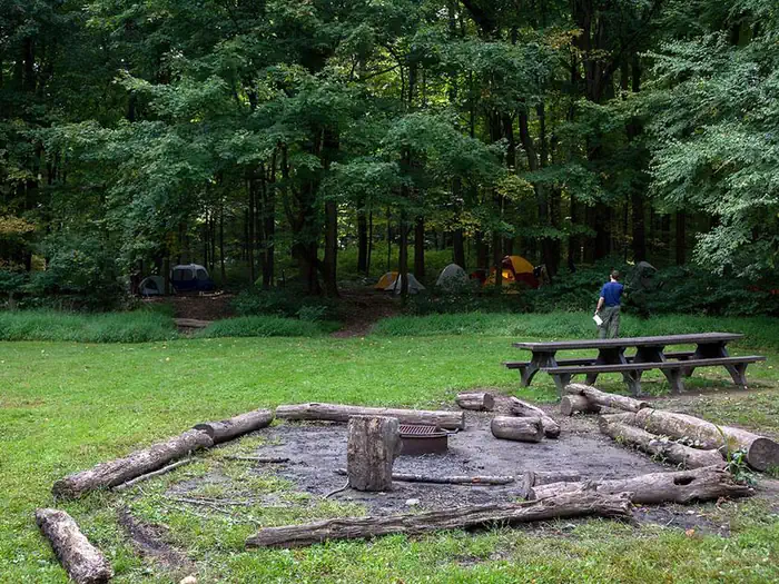 Poplar Grove Youth Group Campground — Metal fire ring/grill surrounded by logs and picnic tables in open field. Multiple tents visible in the distant woods.