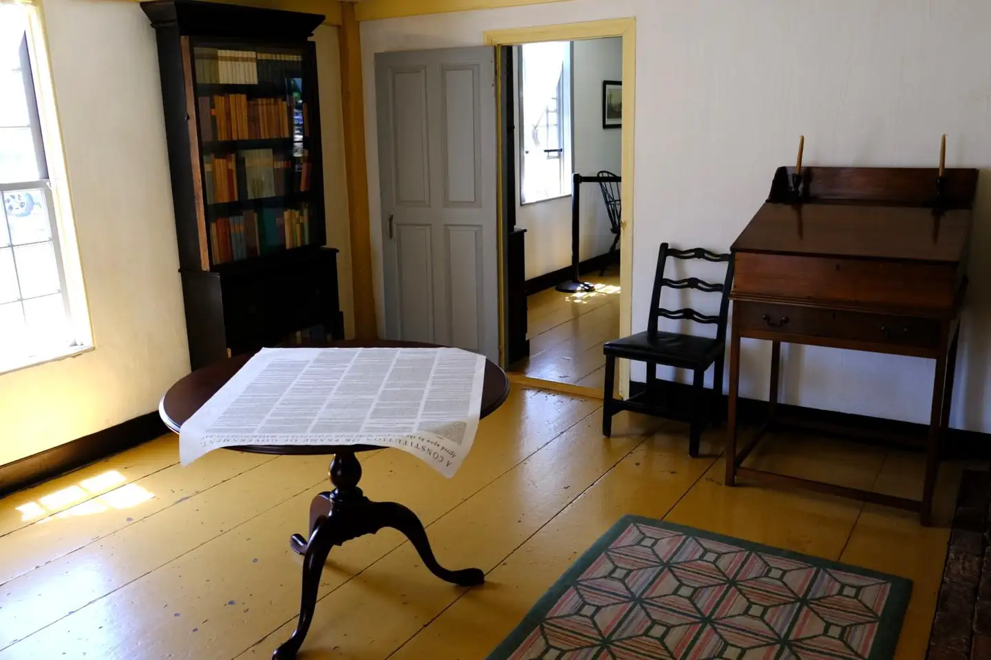 Adams National Historical Park — A room with painted yellow floor, circular table in the middle with large broadside on top, standing 18th-century style desk and chair on the right, and bookcase and window on the left. 