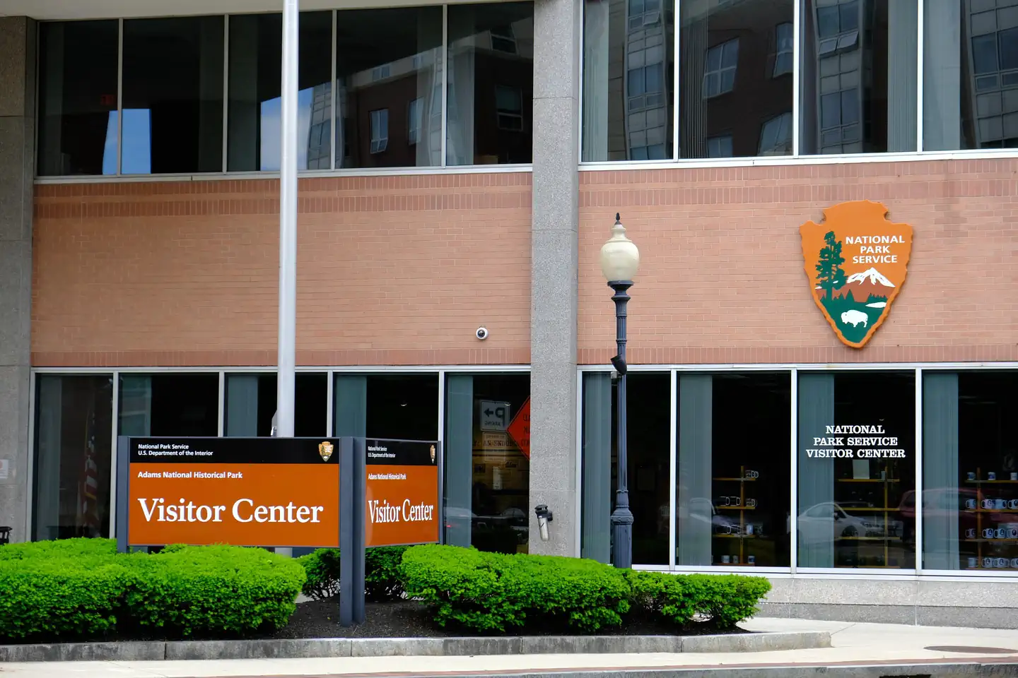 Adams National Historical Park — A sign that reads "Visitor Center" stands in front of a brick and glass office building with large windows. A National Park Service arrowhead hangs from the building. 