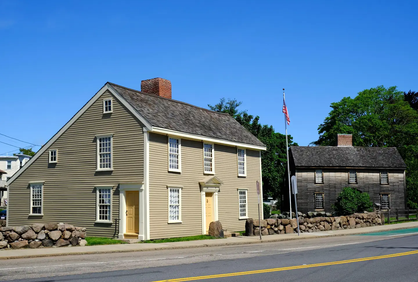 Adams National Historical Park — Two New England "saltbox" style homes sit next to each other. Home on left has gray siding, white trim, and beige doors. Home on right has brown wooden siding and is surrounded by trees and shrubs. Between the buildin