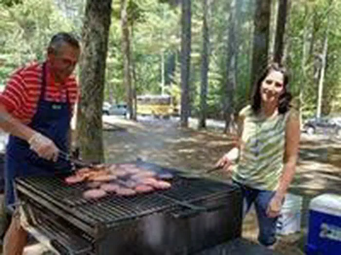West Hill Dam — Two adults turning burgers on a large group-size park provided grill with wooded forested surrounded parking lot in background with cars and school bus.