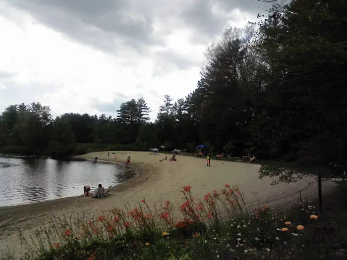 West Hill Dam — View looking down long sandy beach full of visitors on the sand, in the water or along the shore line lawn on picnic tables.