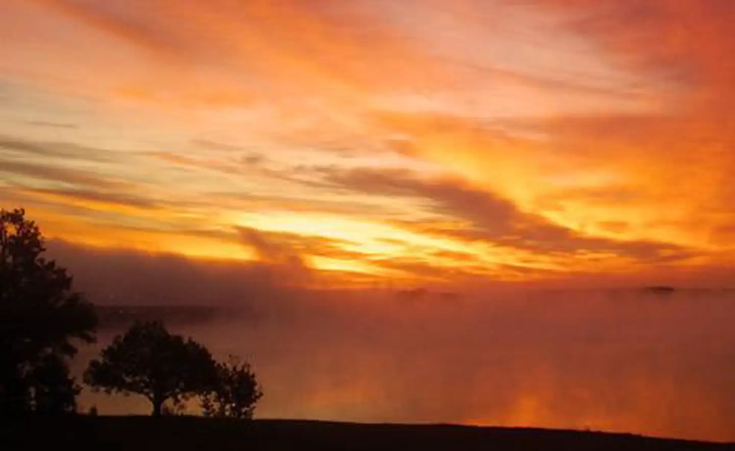 John H. Kerr Dam and Reservoir — This is a picture of the sun rising across the lake. The sky is orange and yellow and there is fog rising off the lake. 