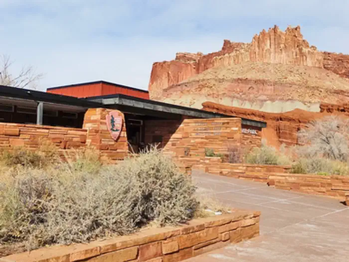 Capitol Reef National Park — A brown stone building. The NPS Arrowhead is on the left side of the building. Desert vegetation is in front of the building. Orang-red cliffs rises above the building. They sky is cloudy with patches of blue.