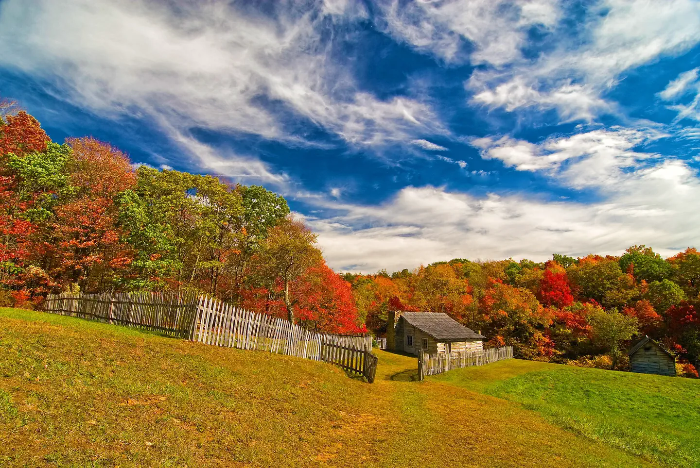Cumberland Gap National Historical Park — A small log cabin and wood fencing in a field, with a forested background of trees with red, green, and orange leaves.