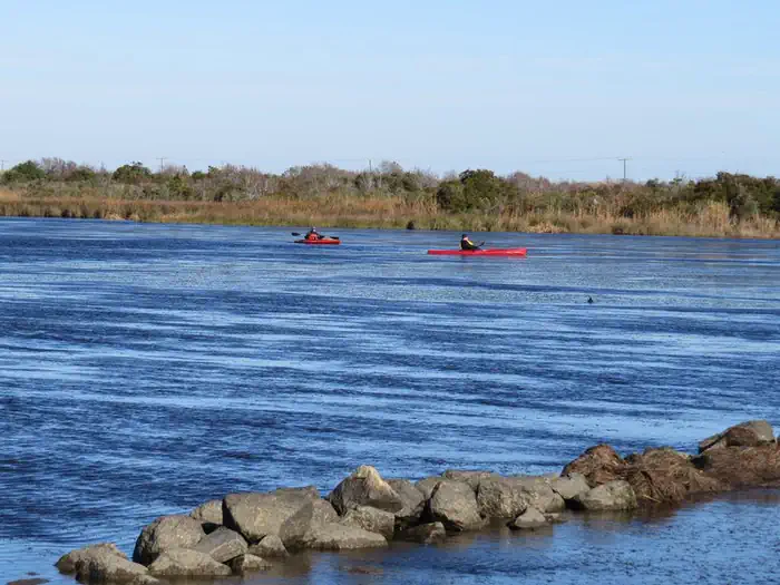 Back Bay National Wildlife Refuge — Two red kayaks float on calm blue water. A line of breakwater rocks line the foreground, with green brown vegetation and trees in the background.