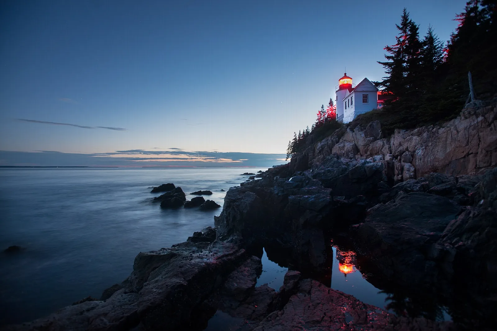 Lighthouse with its lamp lit, perched on dark granite cliffs above calm water at dusk