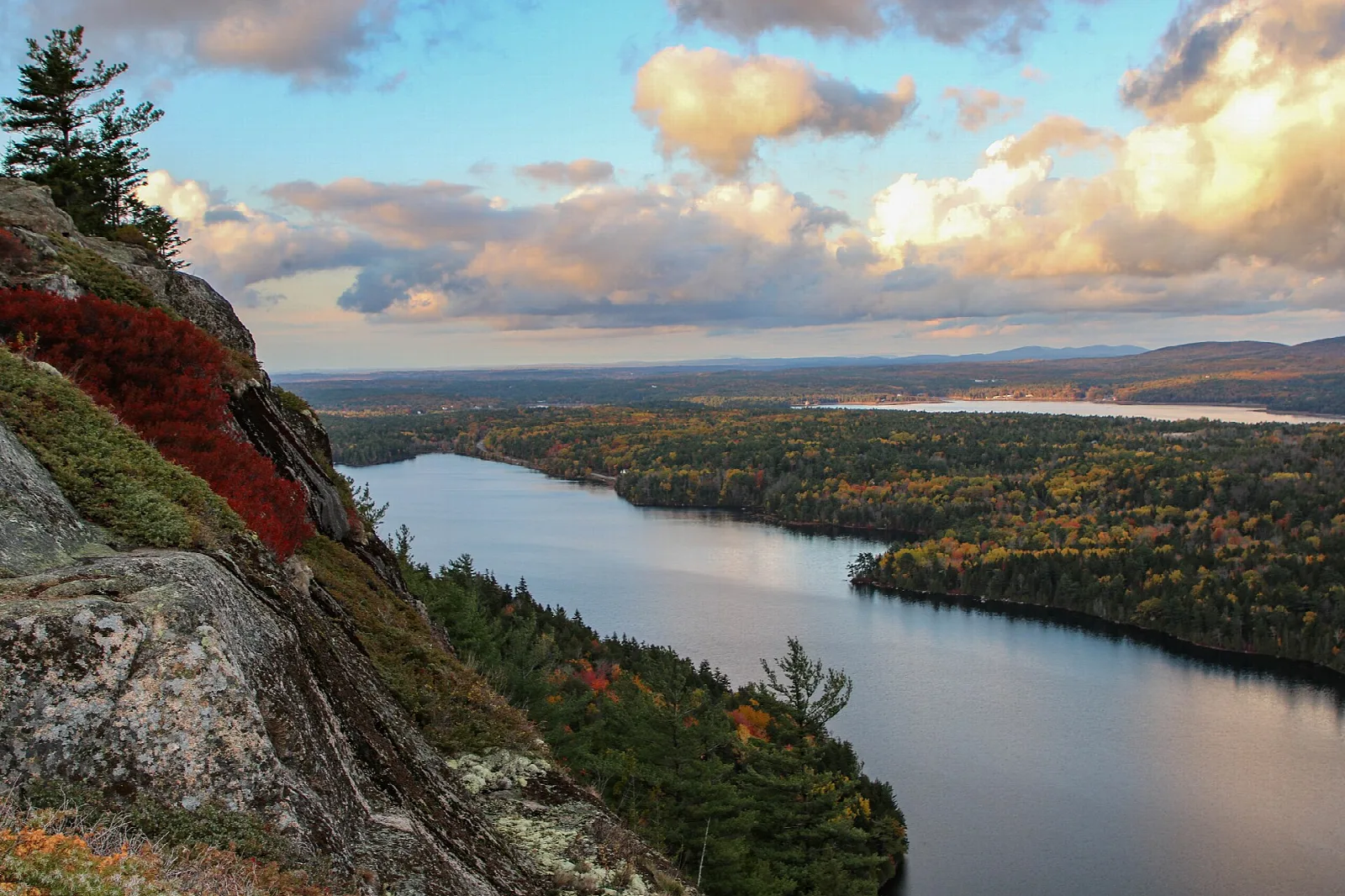 Long lake between forested hills under a partly cloudy sky, seen from a rocky cliff with red foliage