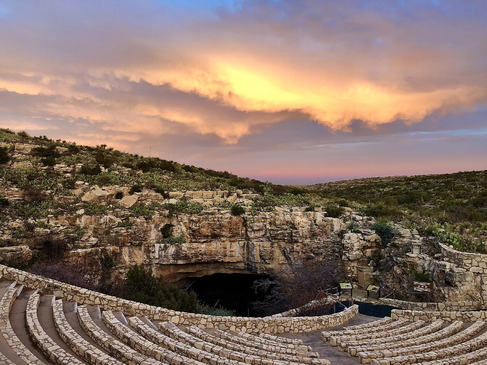 Stone amphitheater seating curving around the cave entrance under a vivid sunset sky