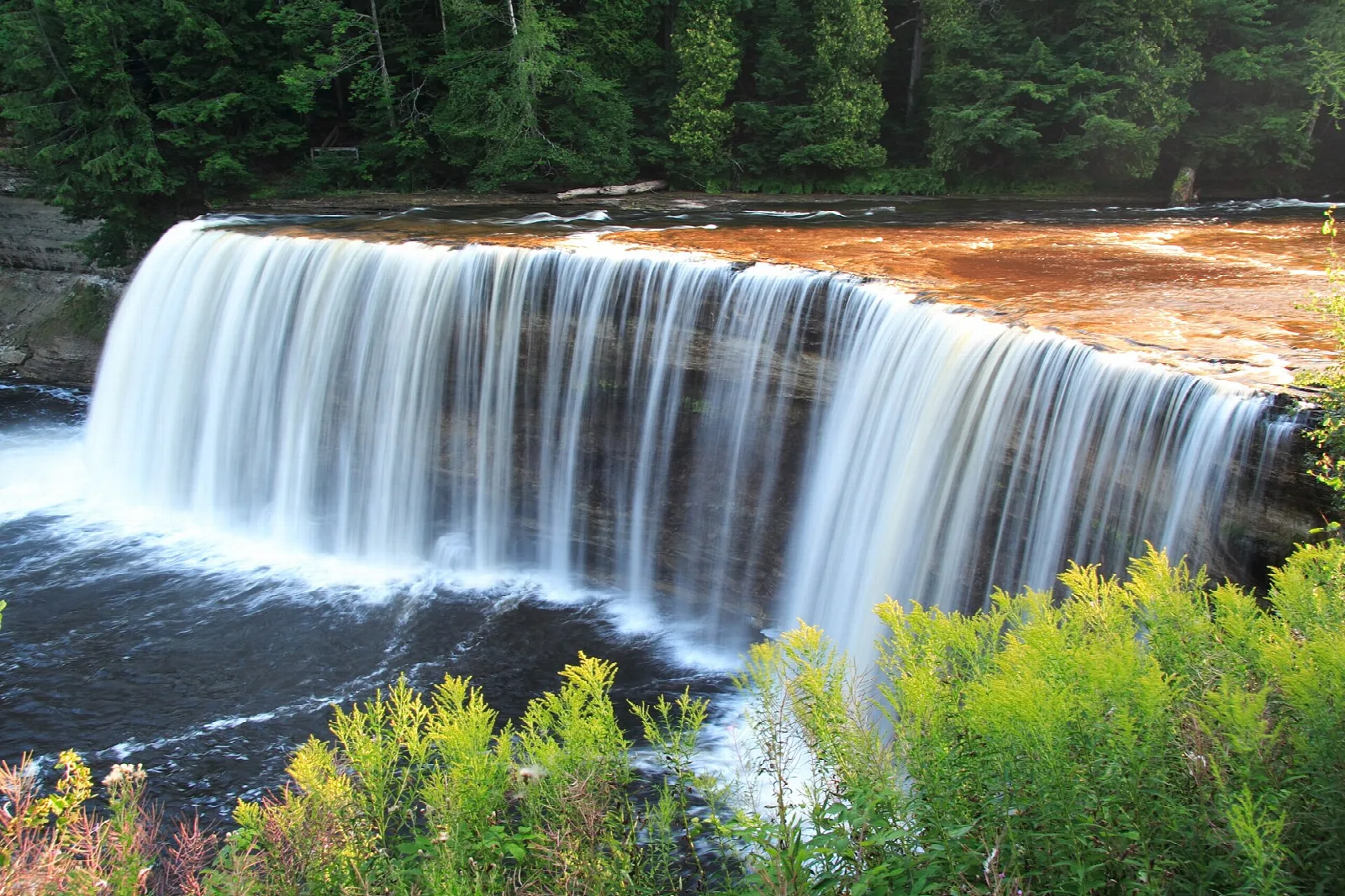 Broad waterfall with white flowing water over a reddish rock ledge