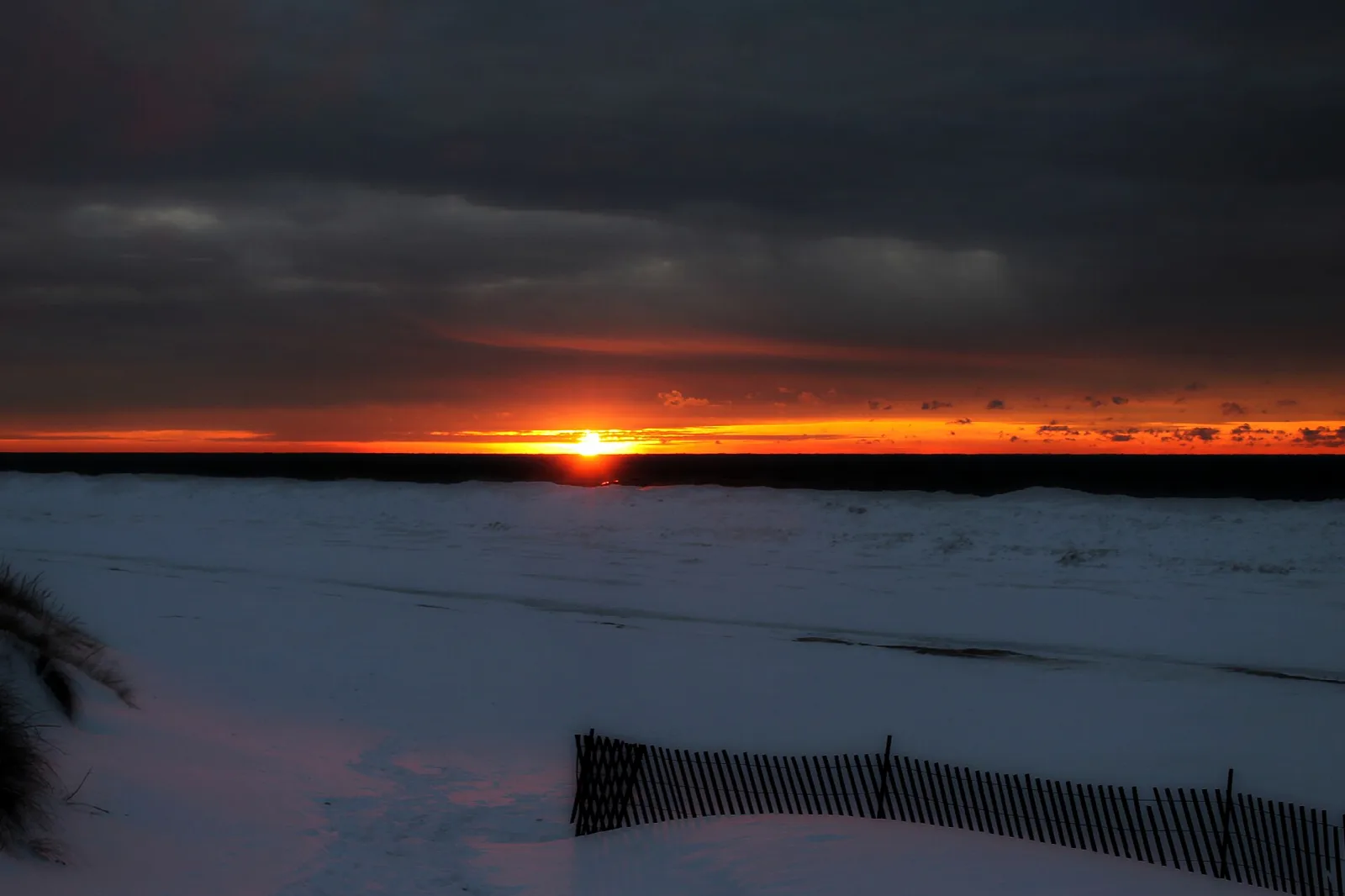 Orange sunset low over a snow-covered shore with a sand fence