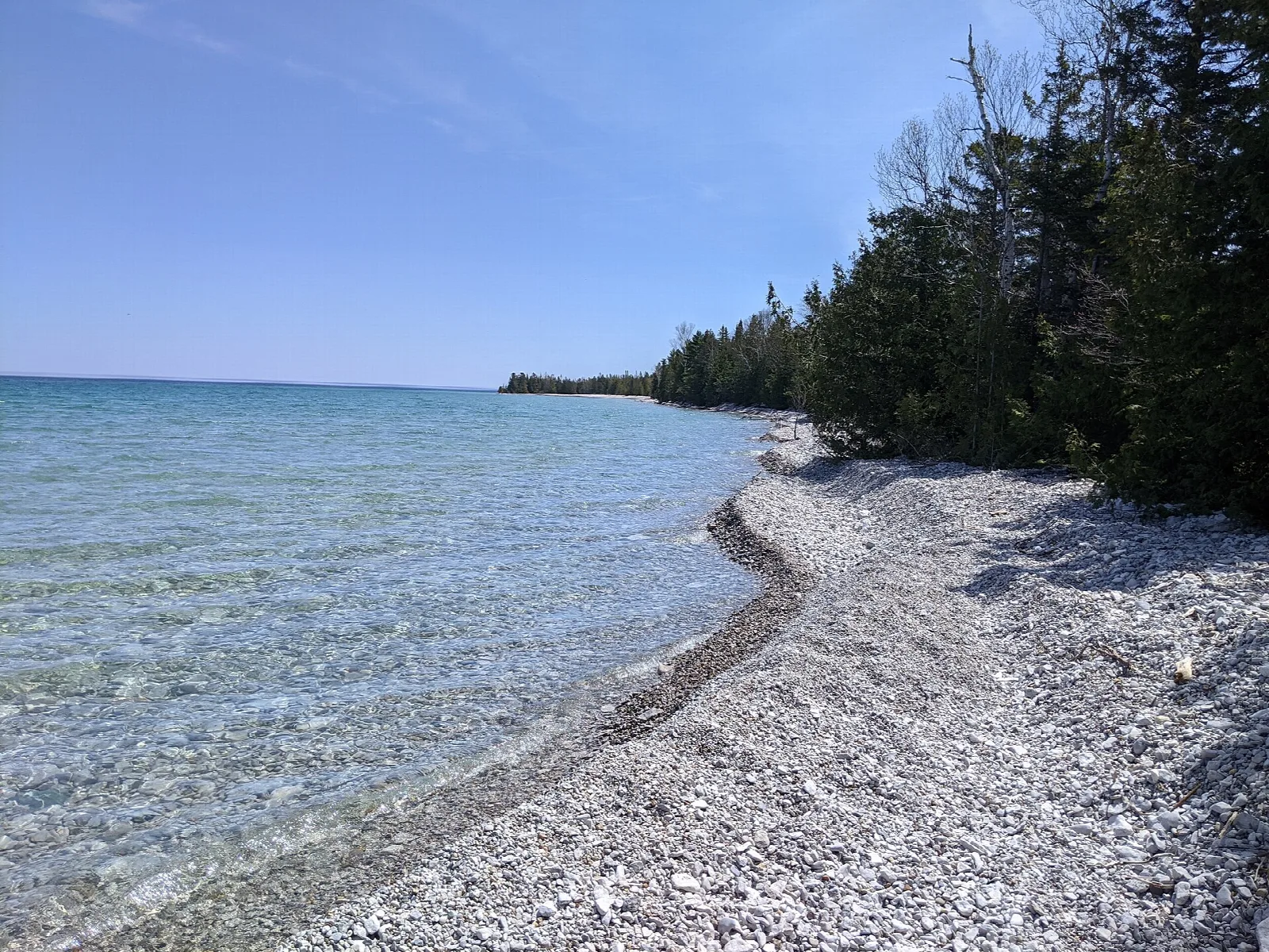 Pale pebble beach along clear blue Lake Huron water with evergreens