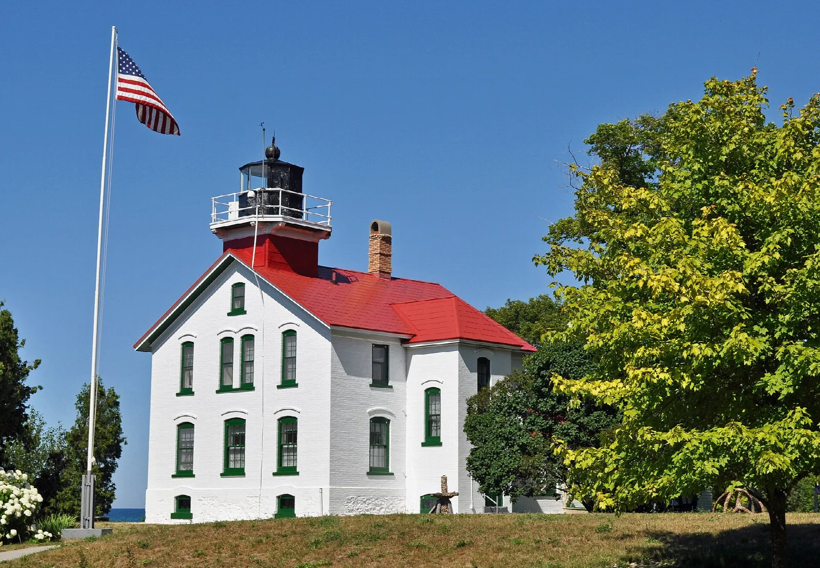 White lighthouse with red roof behind a flagpole and a green tree
