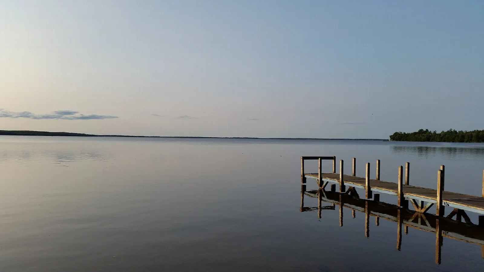 Calm lake with a wooden pier at dusk and a distant tree line