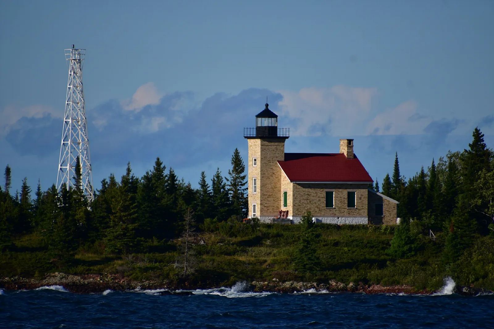 Stone lighthouse with a red roof on a wooded point beside blue water