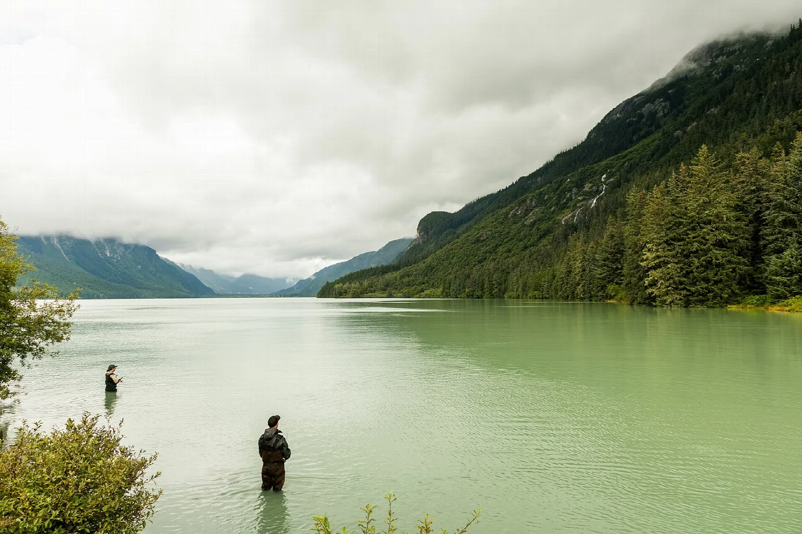 Two anglers wading in shallow green water below a forested mountain